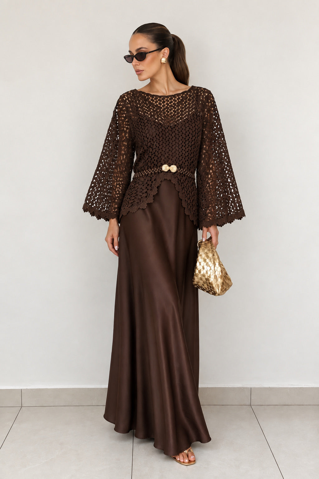 brown two-piece outfit consisting of a long-sleeve crochet top with a gold button detail at the waist and a matching brown satin maxi skirt. She is holding a gold textured clutch and wearing sunglasses, standing against a plain light gray wall.